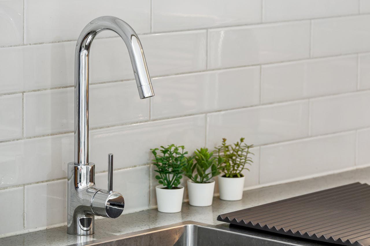 about-02 A sleek kitchen sink with chrome faucet and potted plants on a tiled backsplash.